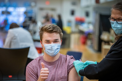 A student wearing a mask gives a thumbs-up signal as a nurse applies a bandage to his arm.