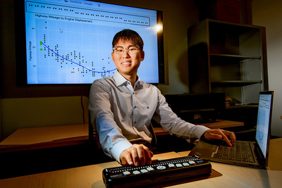 Photo of JooYoung Seo seated at a desk with one hand on a laptop keyboard and the other hand on a refreshable Braille display keyboard, with a screen behind him showing the image of a scatter plot.