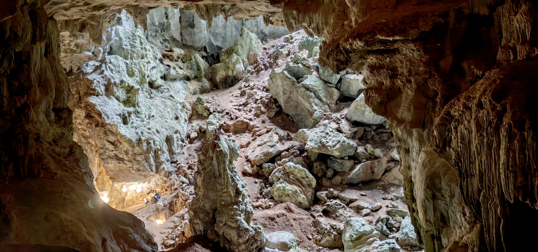 A view of the rocky interior of the cave with light filtering in from above. The team’s bright lights of excavation can be seen in the lower left side of the image.