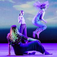 Photo of several dancers onstage, with a seated dancer in the foreground, a dancer leaping in the air in the background, and a projected image of the sky in the background.
