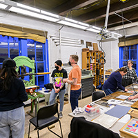 Photo of the Fab Lab’s book arts studio, with some students gathered around the old iron hand press and others around the table reaching for type or print blocks.