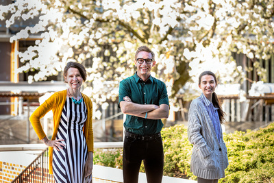Portrait of three, smiling researchers standing outside with a white-flowering tree, bushes and a brick wall in the background.
