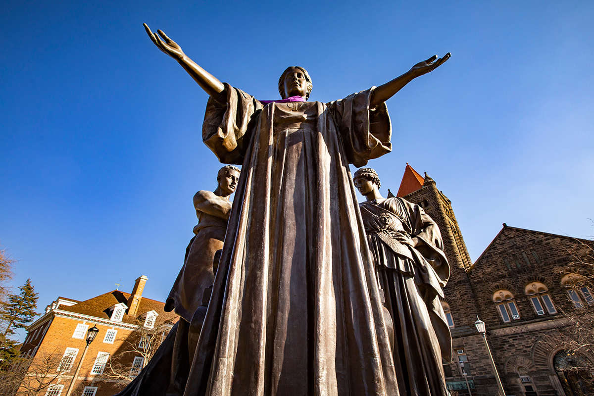 Photo of the Alma Mater statue with outstretched arms, framed against campus buildings and a clear blue sky
