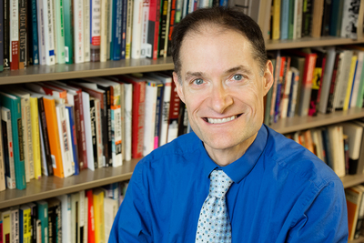 Author Matthew C. Ehrlich wearing a dress shirt and tie standing in front of bookshelves