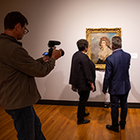 Photo of Maureen Warren and Craig Koslofsky standing in front of a portait in a Krannert Art Museum gallery with a man behind them holding a video camera.