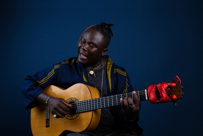 Photo of Okaidja Afroso holding his guitar and singing with his eyes closed.