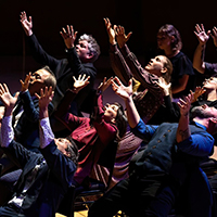 Photo of a group of people leaning back in chairs and singing, with their arms thrown into the air.