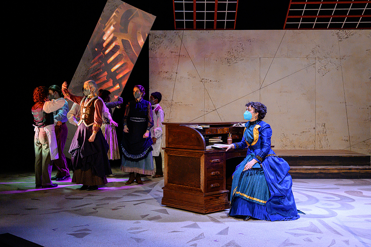 Photo of a woman in a blue Victorian dress seated at a desk onstage, with a group of actors on stage left.