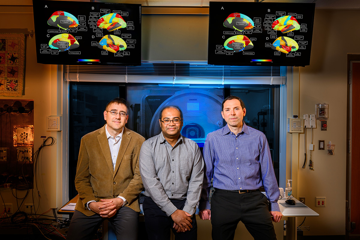The research team in the lab. A screen behind them displays brain regions involved in the new analysis. In the room behind them, an MRI machine.