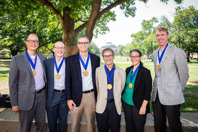 Six people stand outdoors with medallions around their necks