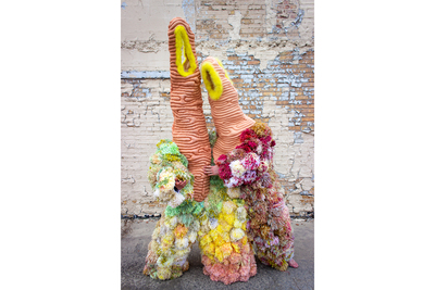 Photo of two people, with only their hands visible, dressed in a large, soft sculpture garment made from pom poms and quilted fabric.