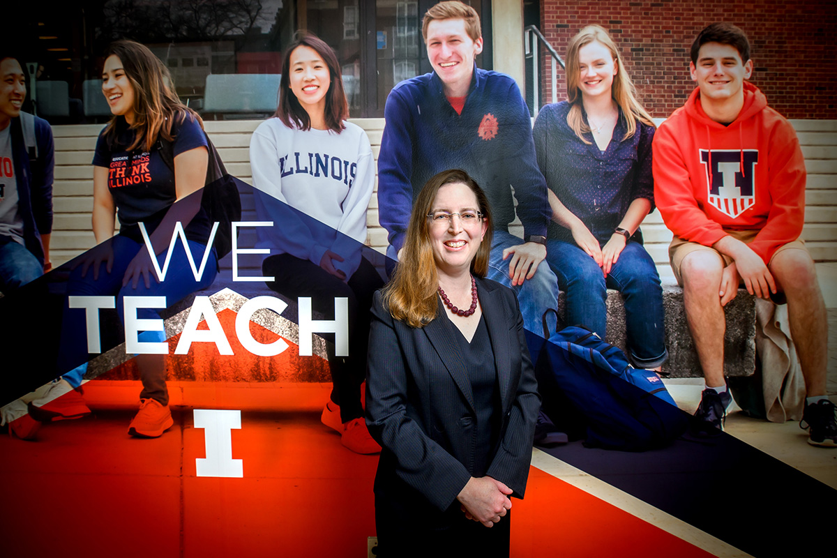 photo of professor Jennifer Delaney standing in front of a mural of several U. of I. students that reads "We teach"