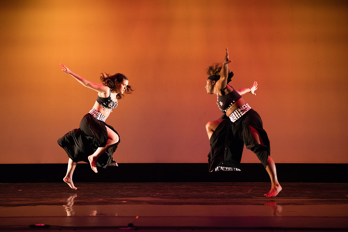 Photo of two dancers dressed in black bra tops and skirts facing each other on a stage and leaping in the air.