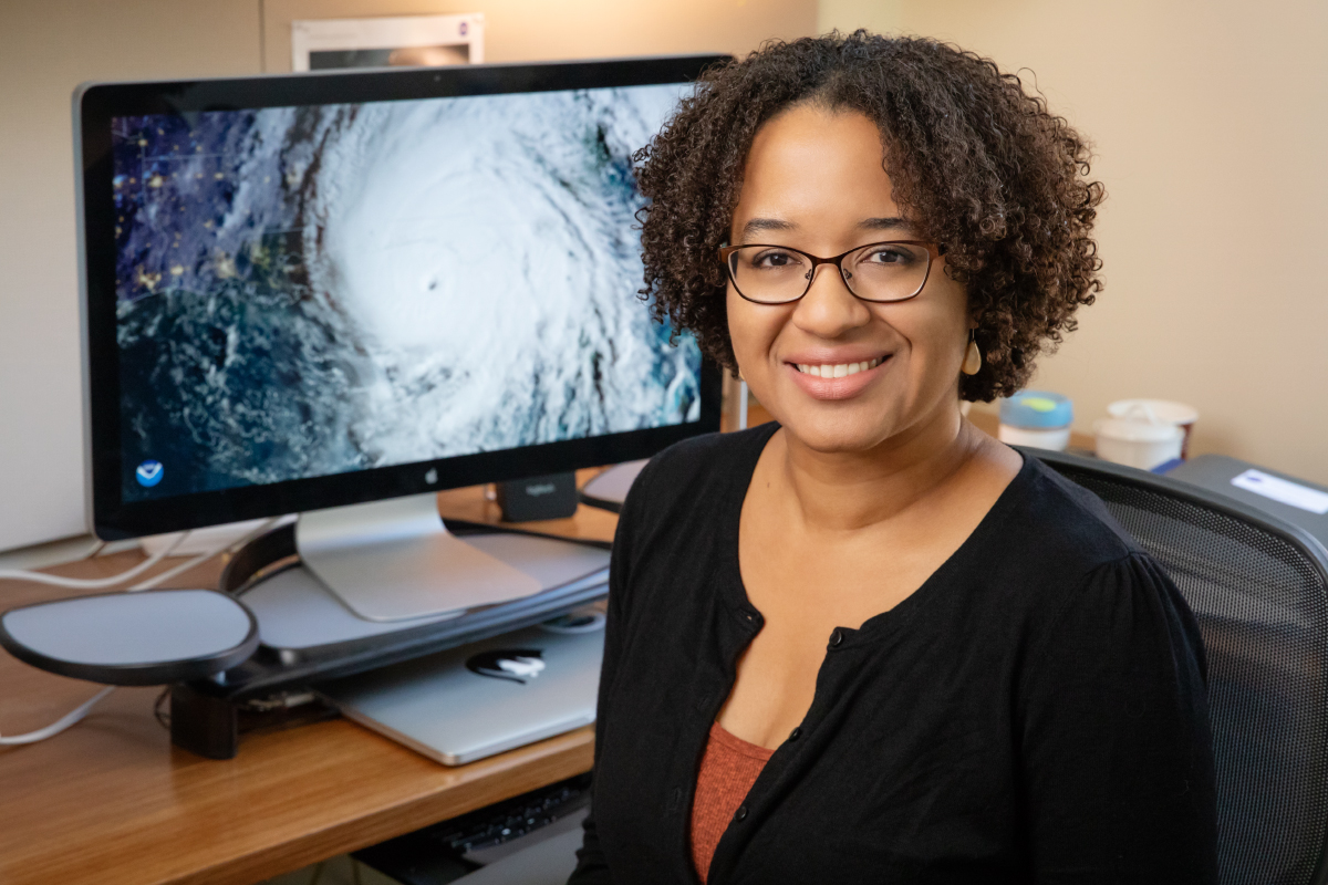 Portrait of professor Deanna Hence, seated, with a computer image of a hurricane in the background