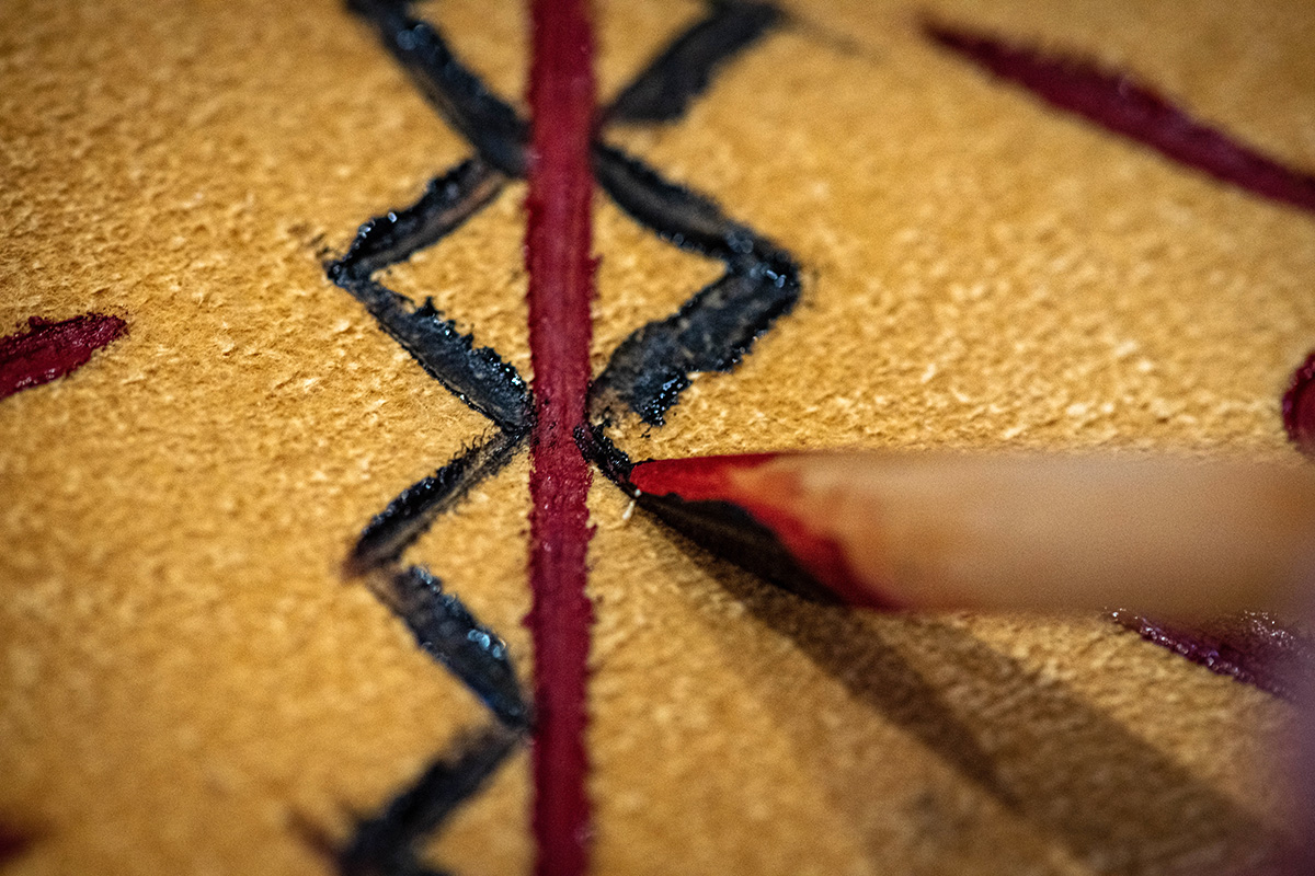 Close-up photo of a wood stylus being used to apply paint to an animal hide, which is being decorated with red and black paint.