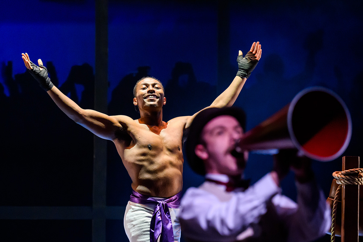 Photo of theatre student Jaylon Muchison in boxing shorts with a purple sash around his waist and his arm raised in the air, smiling, with a man talking through a megaphone in the foreground.