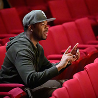 Photo of Tyrone Phillips sitting in theater seats with his hands raised and index fingers pointing, smiling and reacting to action onstage.