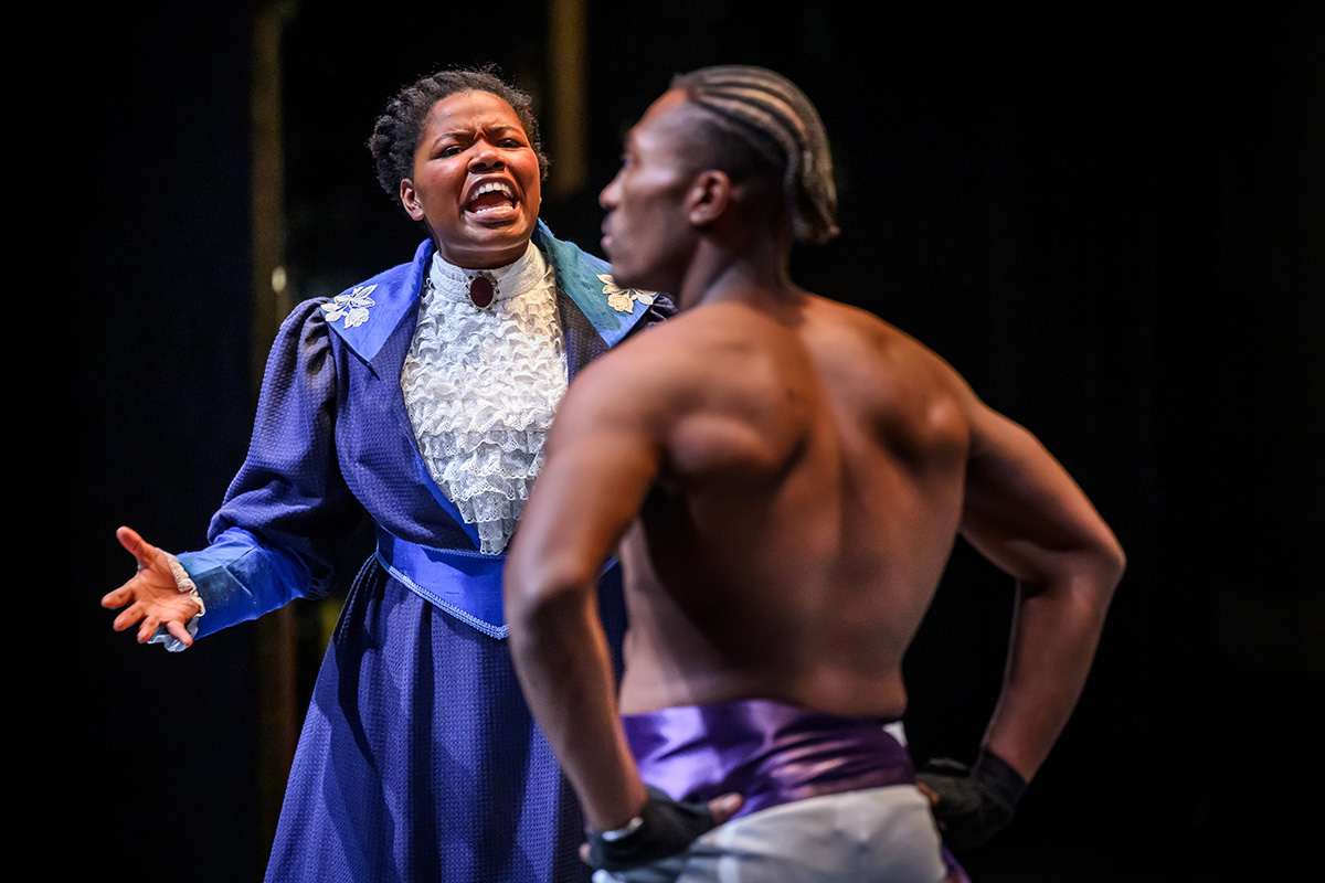 Photo of theatre student Justine Ryan, dressed in a long blue dress and white ruffled shirt, with a brooch at the neck, speaking angrily to Jaylon Muchison, who is in the foreground, seen from the back and wearing boxing shorts.