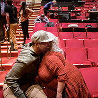 Photo of Tyrone Phillips sitting in theater seats and hugging a woman in an orange sweater whose face is not visible.