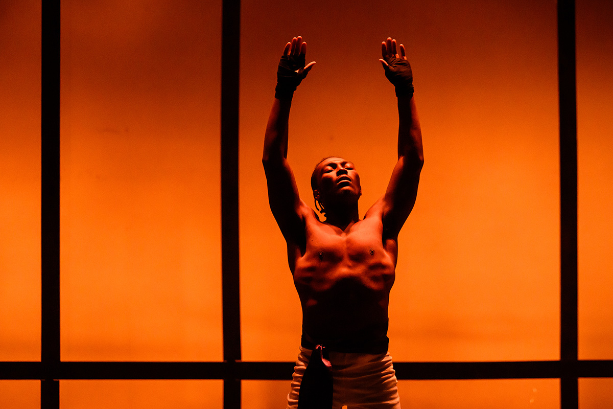 Photo of theatre student Jaylon Muchison onstage in boxing pants with his arms raised and eyes closed, in front of a background lit with orange.