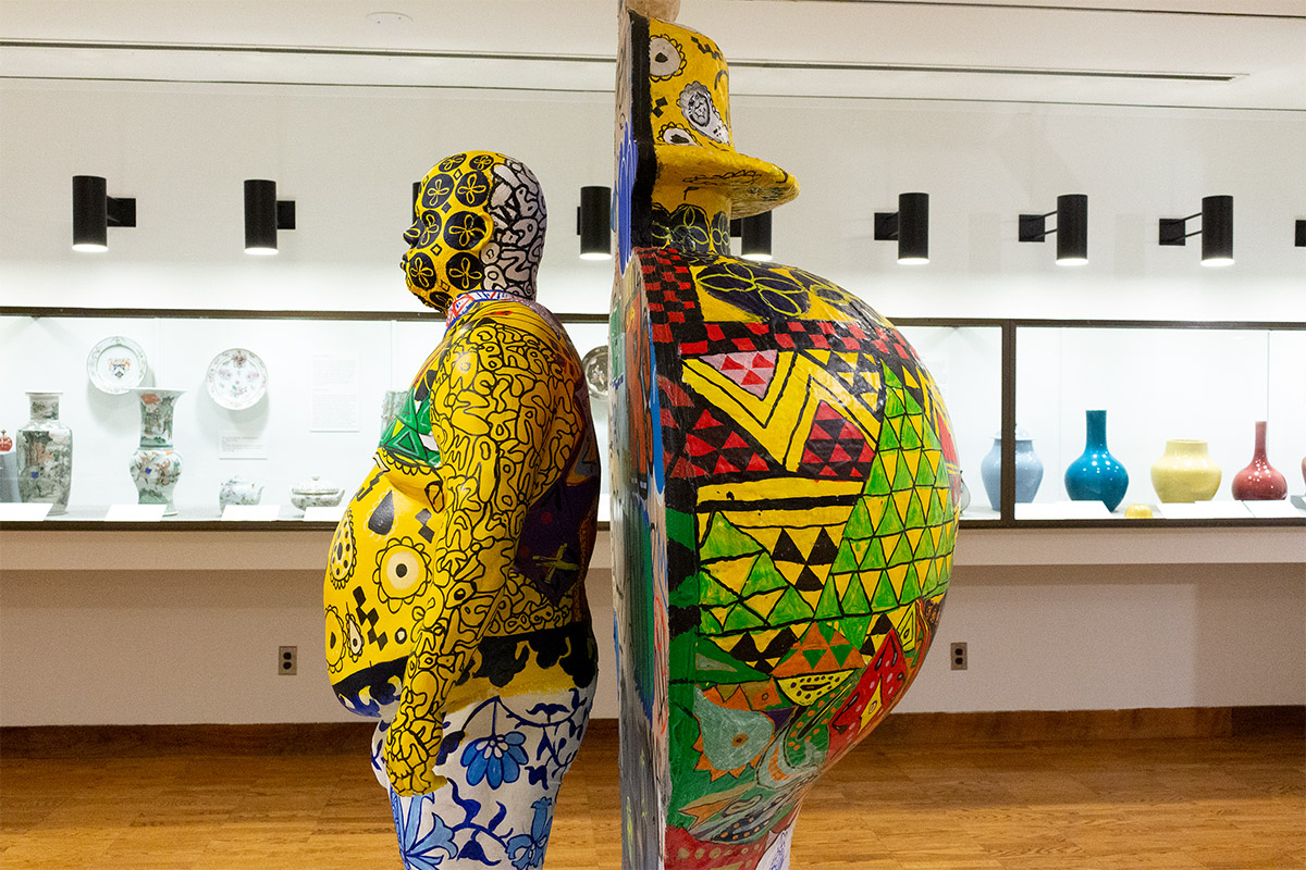 Photo from the side of a sculpture featuring a life-sized cast of a body and a large vase sheared in half. Both are decorated with vibrant colors and patterns.