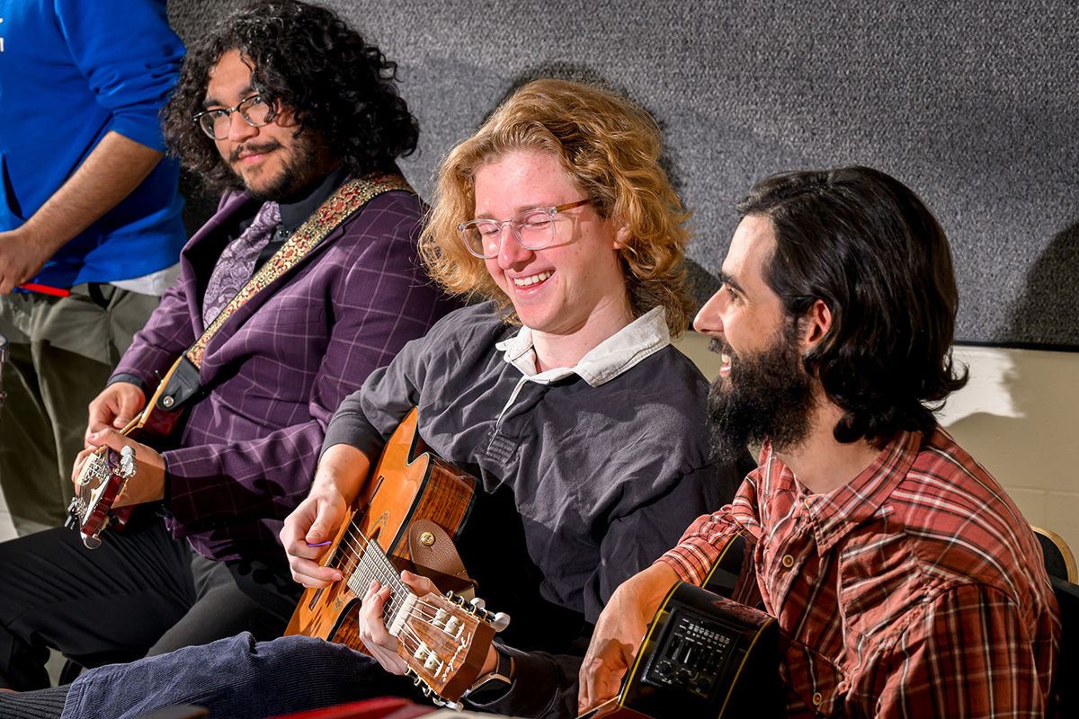 Photo of three guitar players seated next to one another and smiling while they play.