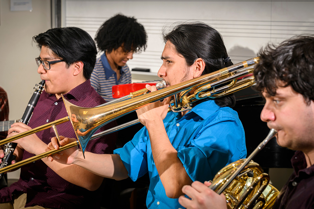 Photo of several musicians playing their instruments -- clarinet, trombone and French horn, with a pianist in the background.