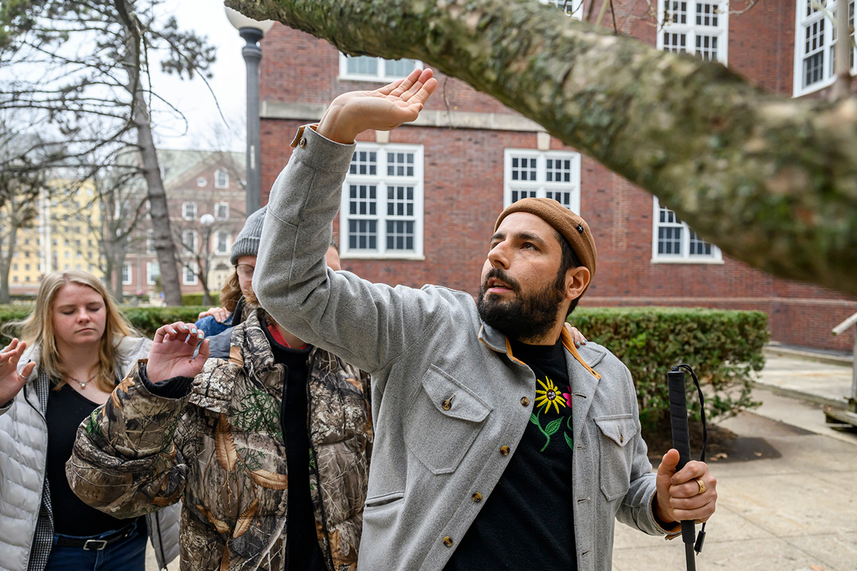 Photo of artist Carmen Papalia raising his arm while warning the line of people following him about a low branch.