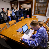 Photo of a group of people holding onto each other’s shoulders and filing past a student sitting at a table with a computer and notebook in front of her.
