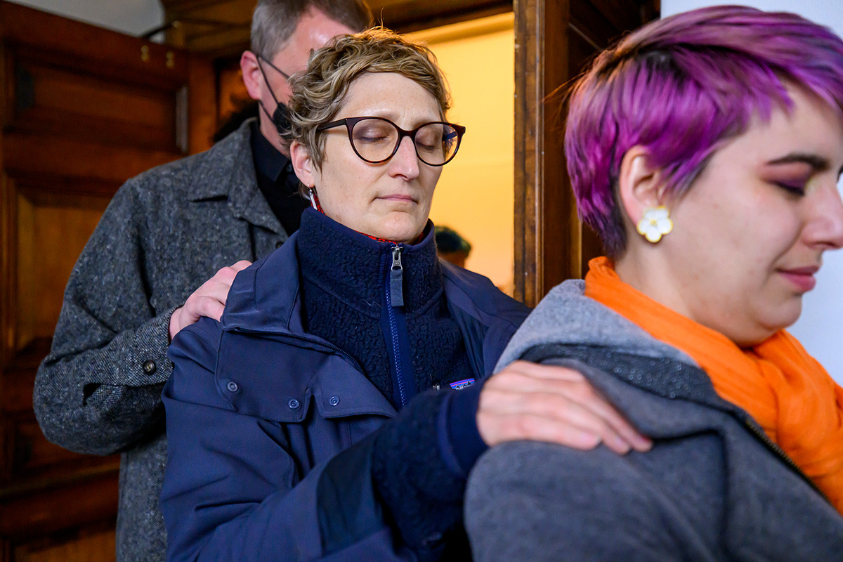 Photo of Annie Adams, with short hair, glasses and wearing a blue coat and fleece, walks in a single-file line with eyes closed, her hand on the shoulder of a woman in front of her with purple hair and an orange scarf.