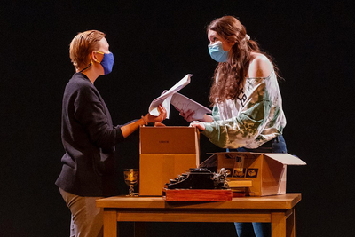 A theatre professor and a student actor perform on stage with a table with a box and typewriter between them.