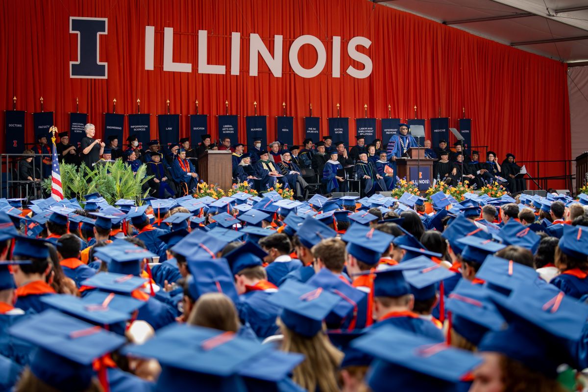 Photo of a crowd of graduates sitting with Chancellor Jones speaking at a podium, other people in graduation regalia seated on a stage and the Block I and "ILLINOIS" across an orange backdrop.