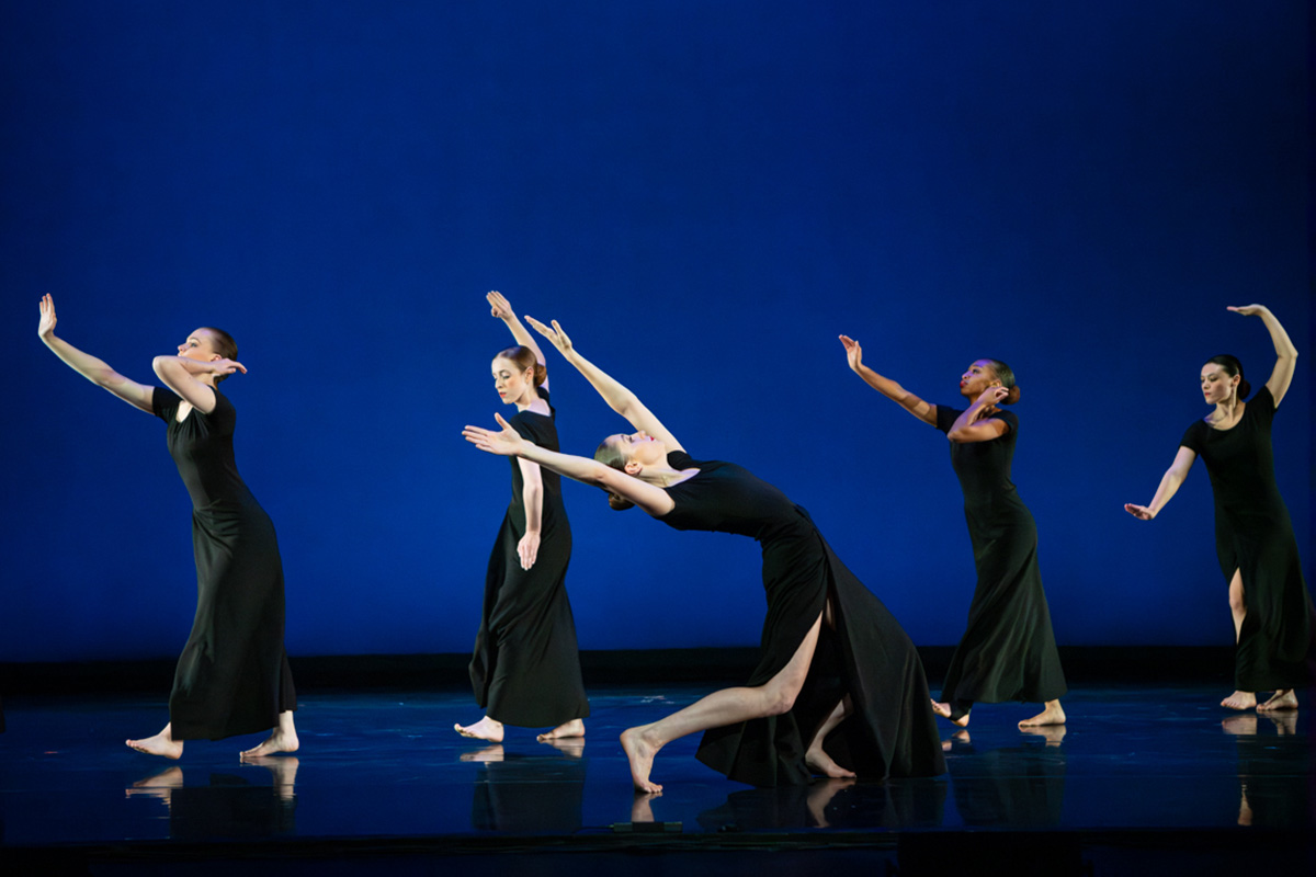 Photo of dancers in black dresses with one in the foreground leaning back with her torso and arms, and four others in the background facing the opposite direction.