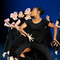 Photo of dancers in black dresses moving in unison with arms and right legs bent as they move forward on stage.