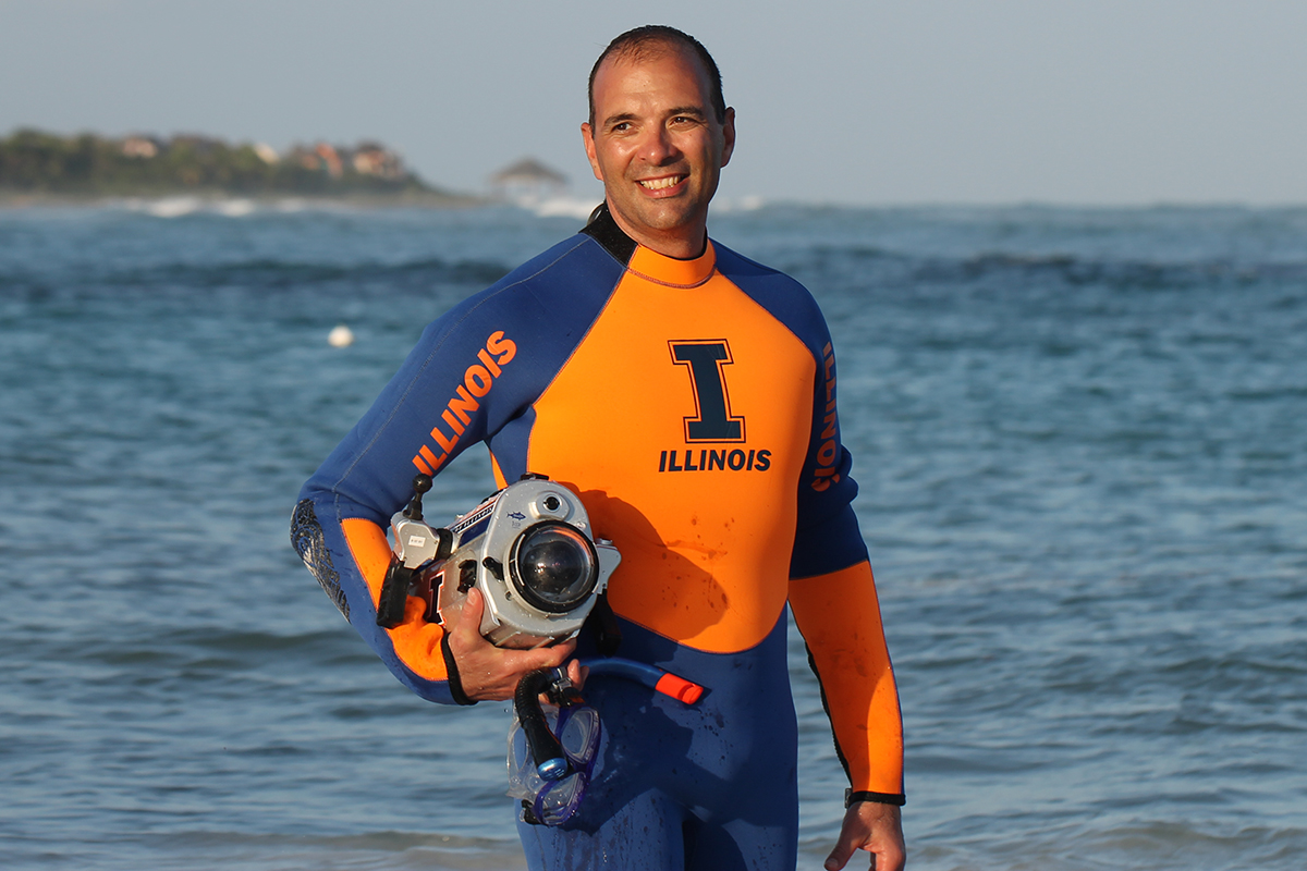 Researcher Viktor Gruev standing in front of the ocean wearing an orange and blue U. of I. wetsuit and holding a specialized camera.
