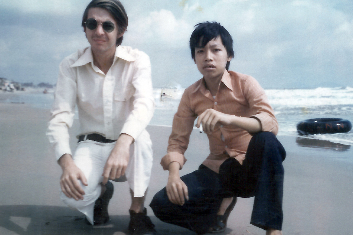 Photo of two men crouching in the sand on a beach, with the ocean behind them. One is a white man wearing white pants, a white buttondown shirt and sunglasses. The other is an Asian man wearing black pants and an orange buttondown shirt and holding a cigarette.