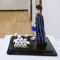Photo of a glass sculpture featuring a masked figure with a pile of toilet paper in front of her.