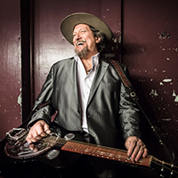 Photo of Jerry Douglas wearing a black leather jacket and hat and holding a guitar on his lap.