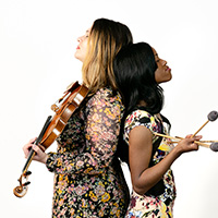Photo of two women standing back to back, one holding a violin and the other holding mallets