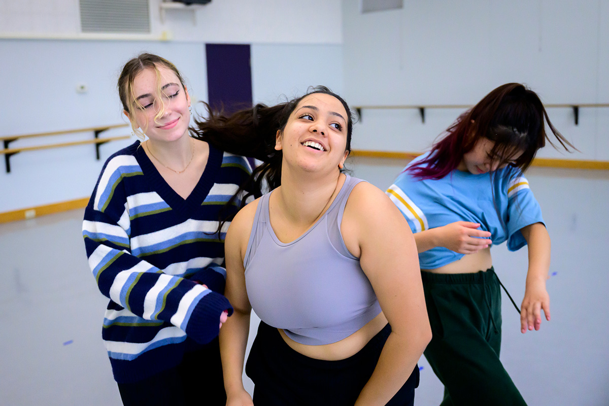 Photo of three women dancing in a studio. One is wearing a blue, black, white and green-striped sweater; one is in a lavendar sports bra; and one has a light blue baseball shirt knotted mid-torso.