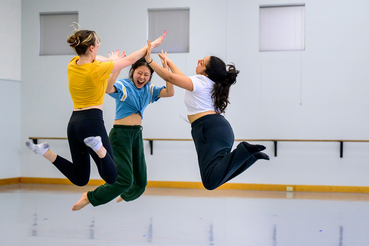 Photo of three dancers in a circle leaping into the air to high-five each other.