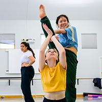 Photo of a dancer swinging her leg over another dancer kneeling before her with her hands up to support the leg of the first dancer, while a third woman watches in the background.