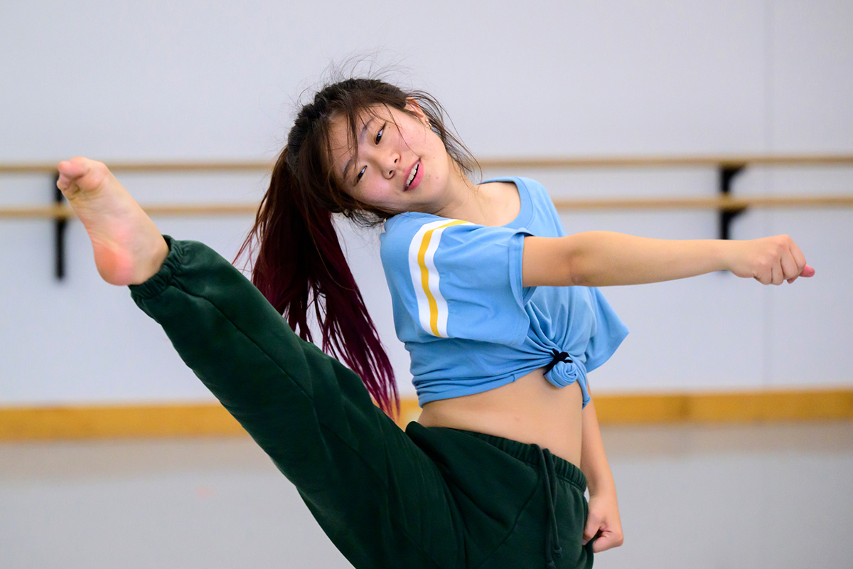 Photo of a dancer with a blue baseball shirt knotted mid-torso kicking her leg up in front of her.