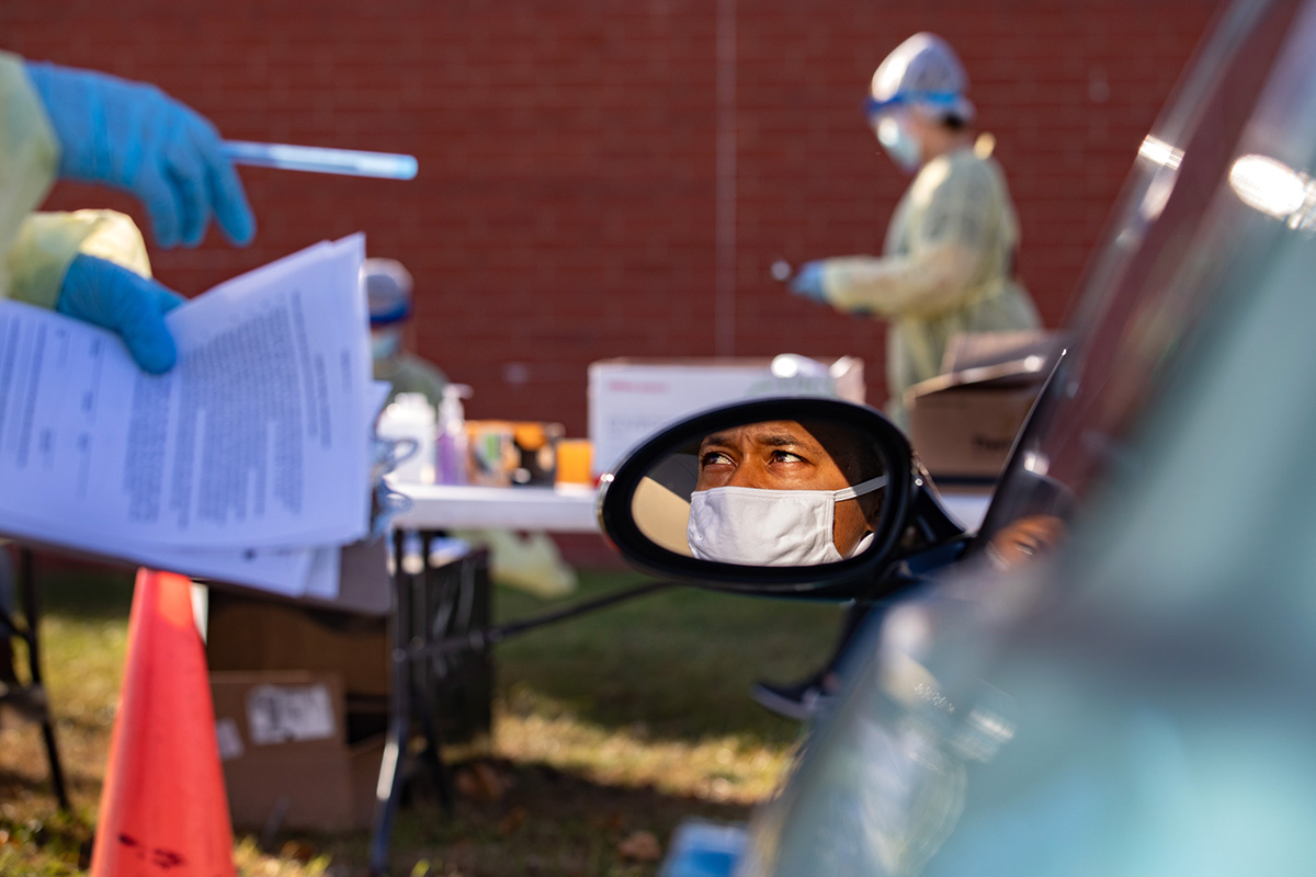 Man wearing face mask shown in car's side view mirror. In front of his car, workers wearing safety gear are preparing to test patients for COVID-19.