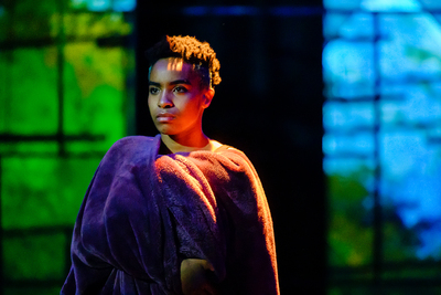 Image of actor Haven Crawley onstage, wrapped in a blanket with green and blue windows in the background.