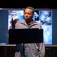 Photo of an actor standing in front of a music stand and gesturing, with a video screen showing abstract images behind him.