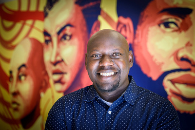 Photo of Moses Okumu standing in front of a colorful mural of men's faces