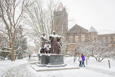 Two people walk in the snow past the Alma Mater sculpture.