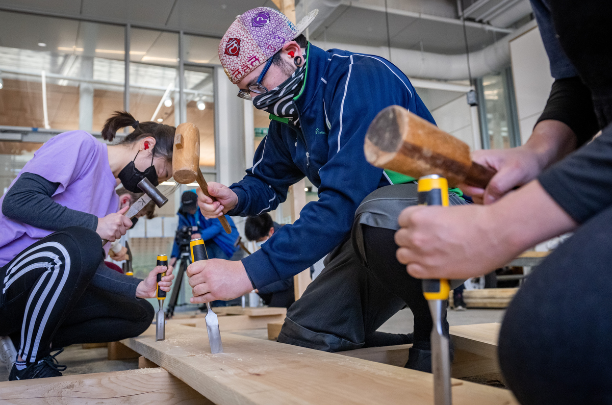 Photo of students kneeling on the floor and working on a plank with chisels and hammers.