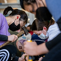Photo of students working along a plank of wood with hammers and chisels.